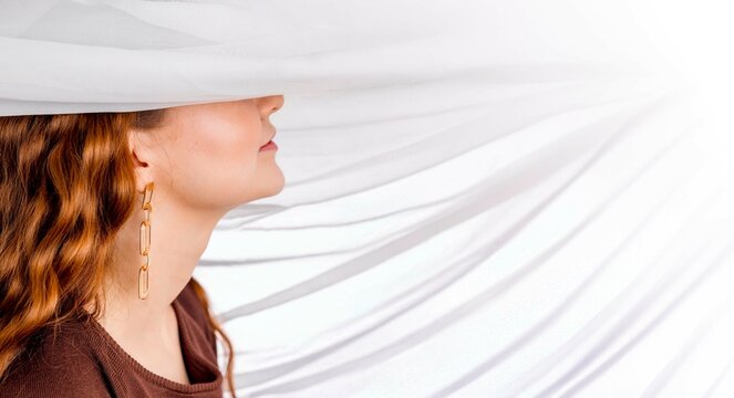 woman with long wavy red hair and golden earrings smiling under white sheer fabric in brown open-back dress, feminine mood in studio, jewelry design, beauty editorial, banner
