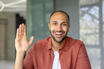 A happy man waves at the camera, smiling broadly and showing off his open palm. He's wearing a...