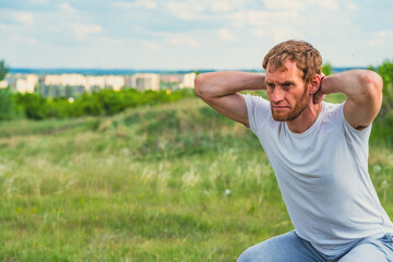 Fototapeta premium A professional sportsman is squatting in a field, with his hands behind his head. Male Performing aerobics exercises, focusing on stretching his shoulders and arms to warm up before a workout.