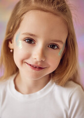 Close-up portrait of a young girl with long blond hair and a gentle smile. She is wearing a white top and has glittery makeup on her face. The background is softly blurred, with pastel tones.