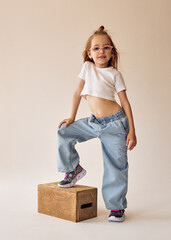 A little mischievous girl with glasses stands in a casual pose with one foot on a wooden box. She's wearing a white crop top and wide jeans. The model with blonde hair poses in a studio on a cyclorama