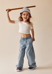 A little girl in a white T-shirt and wide jeans holds a wooden stick above her head and laughs happily. She wears a blue Panama hat, pink glasses. She is playing with a bat joyfully and energetically.