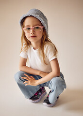 A thoughtful child in a light blue Panama hat, pink glasses, a white T-shirt and jeans sits on her haunches. Her sneakers are decorated with a print of stars and bright contrasting laces