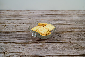 A clear glass bowl holds an assortment of crackers arranged neatly on a rustic wooden surface. The setting suggests a casual snack time at home, perfect for entertaining