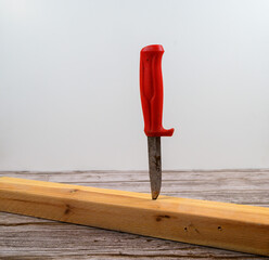 A knife with a bright red handle is embedded firmly into a wooden plank resting on a rustic table. The background is light and neutral, enhancing the focus on the knife