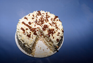 A creamy cake topped with whipped frosting and chocolate shavings sits on a white plate. A slice is cut, indicating it's ready to be enjoyed at a festive gathering