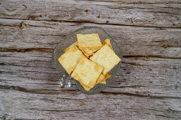 A clear glass bowl holds an assortment of crackers arranged neatly on a rustic wooden surface. The setting suggests a casual snack time at home, perfect for entertaining