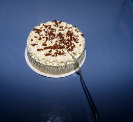 A delicious chocolate cream cake topped with chocolate shavings sits on a circular white platter. The cake is ready to be served at a festive birthday celebration on a blue tablecloth