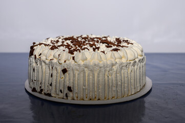 A beautifully decorated layered cream cake sits on a table with rich chocolate shavings on top. The cake features a smooth white frosting and an inviting appearance