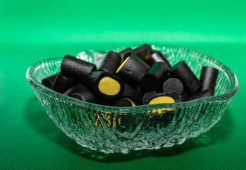 A glass bowl filled with black licorice candies featuring vibrant yellow centers is displayed against a vivid green background. These treats bring a playful touch to the gathering