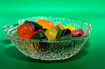 A clear decorative bowl holds a variety of colorful jelly candies, including orange, yellow, green, and red. The vibrant colors contrast against a bright green backdrop