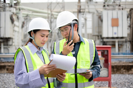 Two engineers work together at power substation with safety helmets and vests while staff communicate and review documents ensuring safe and efficient operation