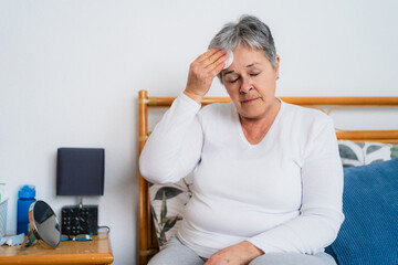 Senior woman experiencing hot flashes or fever, using cotton pad to wipe sweat from forehead while sitting on bed. Skin care