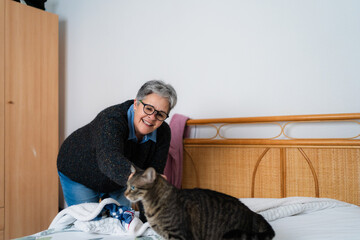Smiling senior woman making the bed in her bedroom while playing with her tabby cat