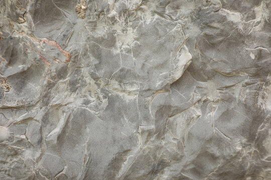 Natural stone surface texture with intricate patterns and fine cracks. The image captures the details of weathered gray rock, showcasing layers, mineral veins, and subtle beige and reddish tones. 