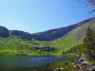 A mountain lake is bordered by rocky slopes and green hills under a clear sky