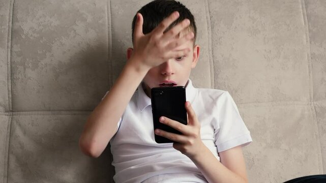 boy 9-11 years old sitting on the couch looks at the smartphone screen in surprise and covers his mouth in horror at what he sees, obscene content for children and teenagers