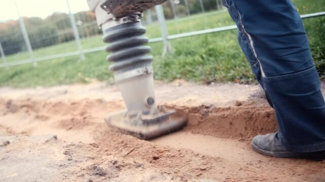 Close-up of a vibrating plate compactor being used by a worker to compact soil or sand at a construction site. The worker's legs in workwear are visible next to the equipment. Concept of groundwork