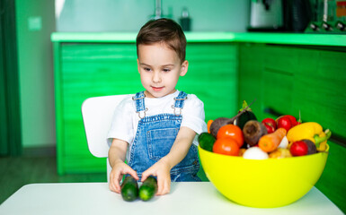 Kid loves fresh veggies!. A young child sits at a white table with cucumbers, surrounded by...
