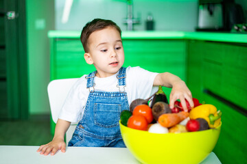 Fototapeta premium Child enjoys colorful fruits in kitchen. A young boy dressed in overalls selects fresh fruits from a bright bowl in a modern kitchen filled with green accents.