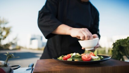 Asian chef adding ingredients to plate and seasoning outdoor. Man in uniform garnishing salad on rooftop. Fresh vegetables and spices on table. Urban background, creating culinary atmosphere.
