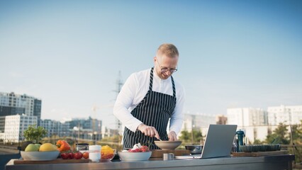Chef checks recipe instructions on a laptop while preparing fresh ingredients. Outdoor rooftop...