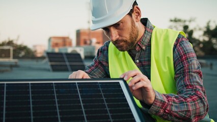 Male engineer in safety gear inspects a solar panel carefully, ensuring its proper alignment and...