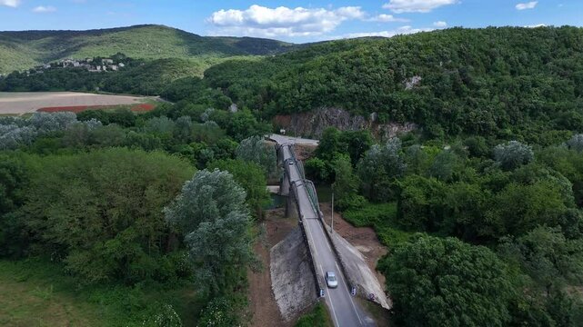 Drone shot gradually approaching a narrow steel bridge as a car crosses it, rising in altitude while tilting the camera downward to reveal the lush green valley and surrounding hills