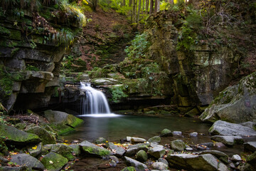 Waterfall in the heart of the Beskydy Mountains, surrounded by moss-covered rocks and lush forest. A long exposure captures the gentle flow of water falling into a tranquil pool in a secluded woodland