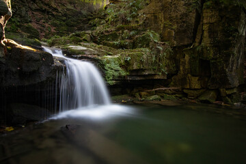 Waterfall in the heart of the Beskydy Mountains, surrounded by moss-covered rocks and lush forest. A long exposure captures the gentle flow of water falling into a tranquil pool in a secluded woodland