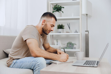 Focused man writing in notebook while sitting comfortably at home. The modern workspace features a...