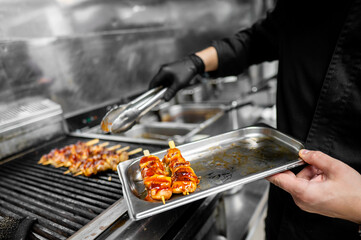 A professional chef grills skewered meat, coated in a glossy sauce, using tongs. The chef holds a...