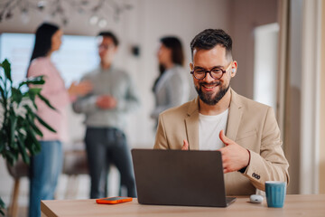 Fototapeta premium Businessman making video call on laptop in office with colleagues discussing in background