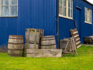 Rustic wood barrels and crates beside a weathered blue building on grass.