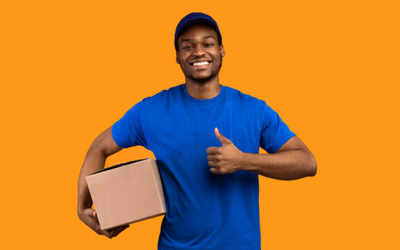 Best Delivery Service Concept. Smiling black male courier wearing blue cap and uniform holding cardboard box and showing thumbs up gesture standing isolated over yellow orange studio background