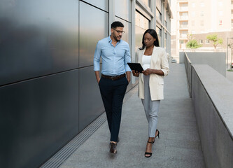 Two Multiethnic business people walking and talking about work outside office using digital tablet