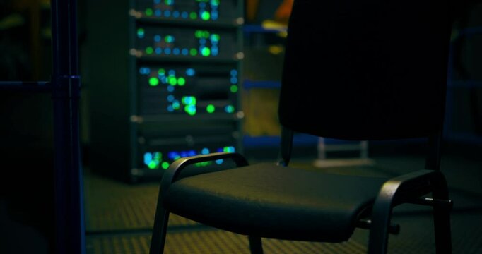 A vacant chair stands in a dimly lit server room surrounded by rows of servers. Colorful lights flash intermittently, creating a mysterious atmosphere in the late hours.