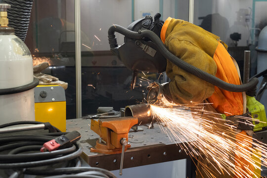 Metalworker grinding pipe with electric tool, producing sparks in a safety-equipped workshop environment. Wearing helmet and flame-resistant suit. &ndash; Metalworking, Industrial Manufacturing