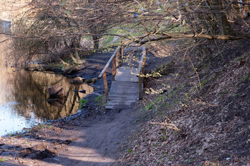 A small arched wooden footbridge made of planks spans a muddy stream flowing down a forested slope into a lake. On the left, the lake and a shoreline; on the right, a steep hill covered with trees. 