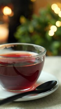 Gentle push-in video of rich translucent red tea in a clear glass cup on a white ceramic saucer, subtle textured white linen, soft focus dark wooden spoon. Warm candle light, leafy branches, bokeh