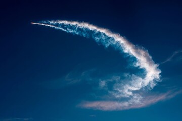 A rocket ascends into the sky, leaving a striking smoke trail against a backdrop of clear blue. The launch occurs during the early evening when the sky is mostly clear