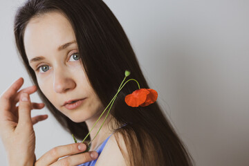 A beautiful brunette young white woman gazes at the viewer, with a red poppy gently resting in her...
