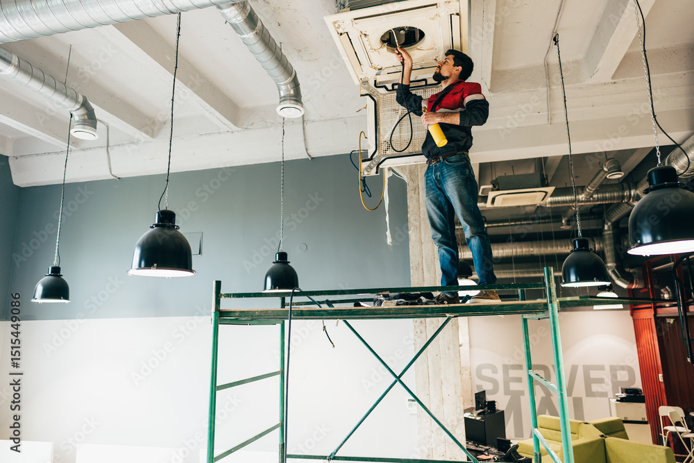 Poster worker performs maintenance on hvac system while standing on scaffolding in an office space during t