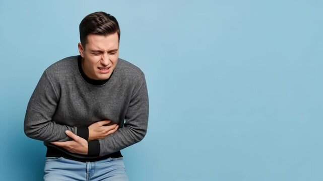 A young man is sitting down, holding his stomach in discomfort. He shows signs of pain on his face as he leans forward slightly in an indoor environment with a blue background