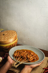 Traditional Indonesian chicken satay with peanut sauce served on a plate. Hands presenting the dish with rustic background and spices.