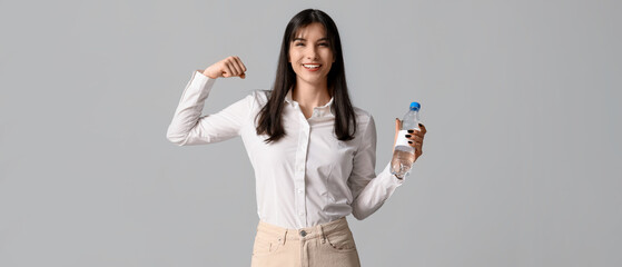 Young Asian woman with bottle of water showing strength on grey background