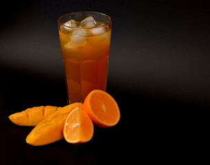 Orange-mango juice in a tall faceted glass with ice on a black background, next to pieces of ripe fruit.