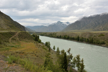 High banks of a wide mountain river flowing through a long valley surrounded by rocks on a cloudy summer day.