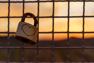 Love padlock with heart engraving on a wire fence at sunset. Symbol of romantic commitment. Blurred background with warm tones. Shot in Pisa, Italy.