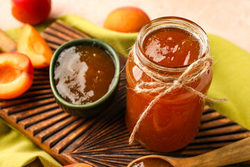 Wooden board with bowl and jar of sweet apricot jam on pink background